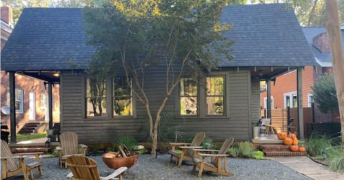 A cozy gray house with large windows, surrounded by trees and a gravel yard with wooden chairs and pumpkins.