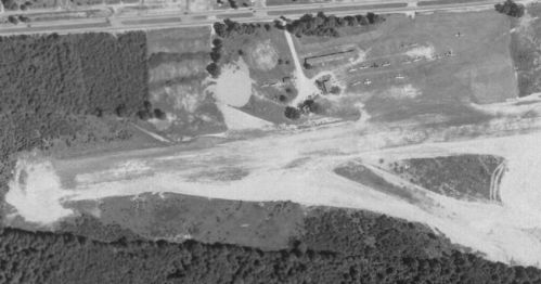 Aerial view of an airstrip surrounded by trees, with several aircraft parked nearby and a building in the background.