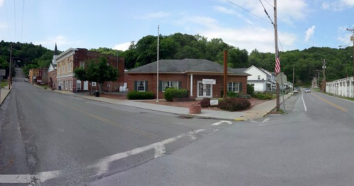 A small town intersection featuring brick buildings, greenery, and a flagpole under a blue sky.