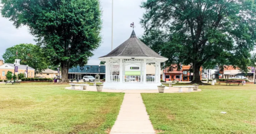 A white gazebo surrounded by green grass and trees, with a pathway leading to it in a small town setting.