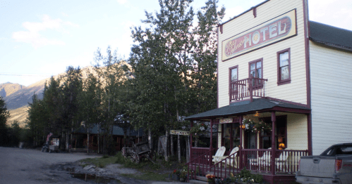 A quaint, vintage hotel surrounded by trees and mountains, with a porch and flower pots in a rustic setting.