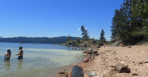 People enjoying a sunny day at a lake beach, with trees and mountains in the background.
