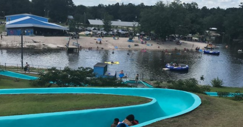 A water park scene featuring a blue waterslide, a sandy beach, and people enjoying boats on a lake.