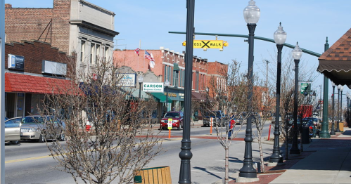 A quiet downtown street lined with shops, trees, and street lamps on a clear day.