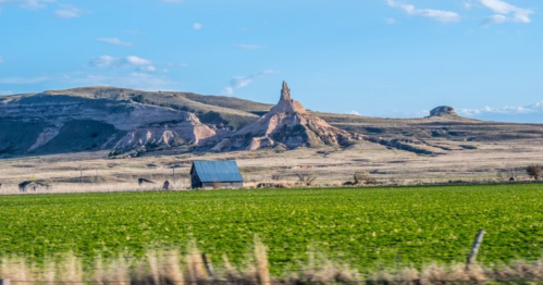 A scenic landscape featuring a green field, a rustic barn, and a prominent rock formation under a blue sky.