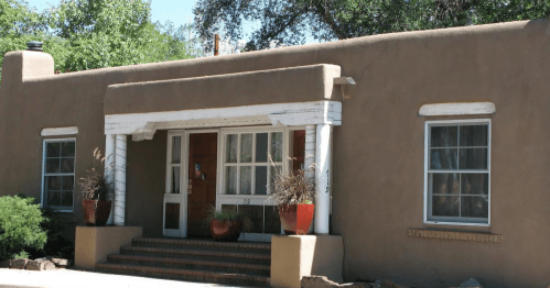 A southwestern-style adobe house with a porch, white trim, and potted plants on either side of the entrance.