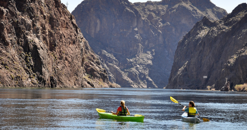 Two people kayaking in a river surrounded by steep, rocky cliffs under a clear blue sky.