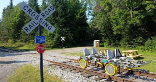 A rail bike parked on train tracks near a stop sign and a railroad crossing sign, surrounded by trees and picnic tables.