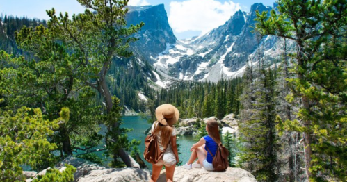 Two women sit on rocks by a lake, gazing at majestic mountains and lush greenery under a bright blue sky.