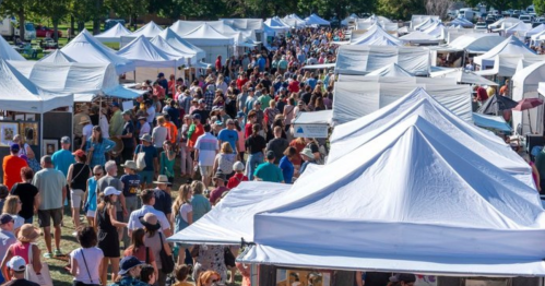 Aerial view of a crowded outdoor market with numerous white tents and a large crowd of people browsing.