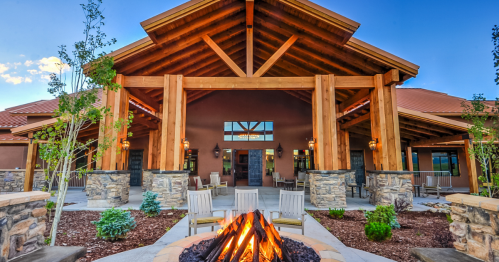 A modern lodge with a wooden structure, stone accents, and a fire pit in the foreground, surrounded by greenery.