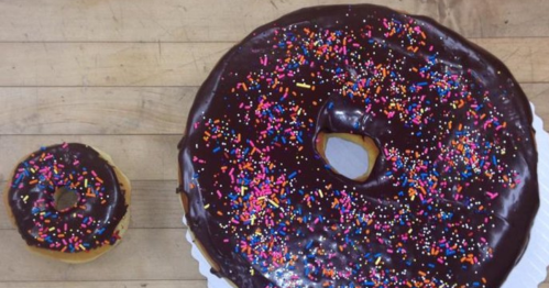 A large chocolate frosted donut with colorful sprinkles next to a smaller donut on a wooden surface.