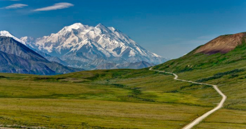 A winding dirt road leads through lush green fields, with majestic snow-capped mountains in the background under a clear blue sky.