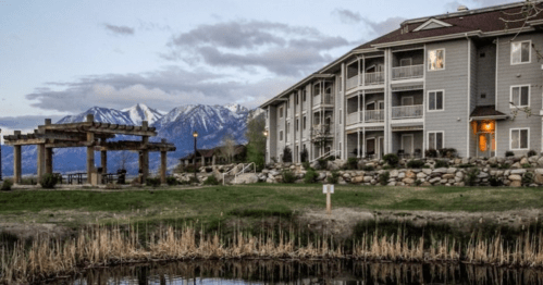 A lakeside view of a modern building with mountains in the background, surrounded by greenery and a calm water reflection.