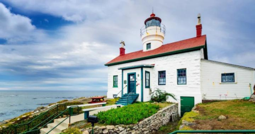 A white lighthouse with a red roof stands by the ocean under a cloudy sky, surrounded by green grass and a stone wall.