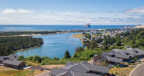 Aerial view of a coastal town with homes, a river, and a rocky outcrop under a clear blue sky.