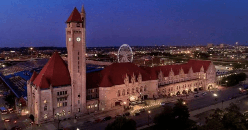 Historic train station at dusk, featuring a clock tower and surrounding city lights, with a Ferris wheel in the background.