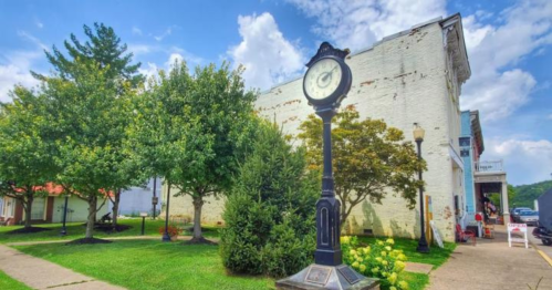 A vintage clock stands in a green park area with trees and a historic building in the background under a blue sky.