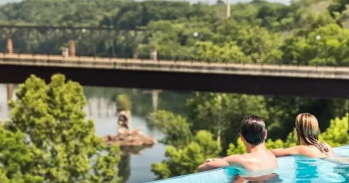 A couple relaxes in an infinity pool overlooking a river and a bridge surrounded by lush greenery.