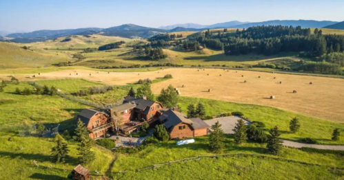 Aerial view of a rustic farmhouse surrounded by green fields and rolling hills under a clear blue sky.