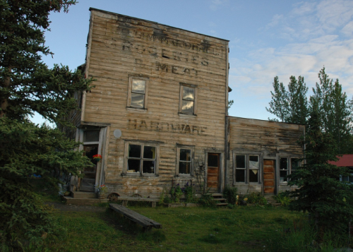 Abandoned wooden building with faded signage, surrounded by greenery and trees under a blue sky.