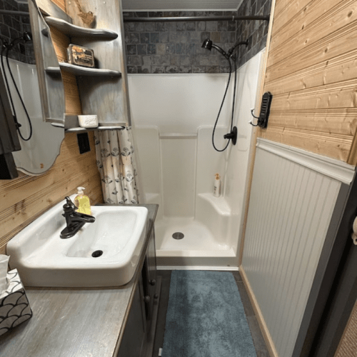 A small bathroom featuring a shower, sink, wooden walls, and a blue rug.