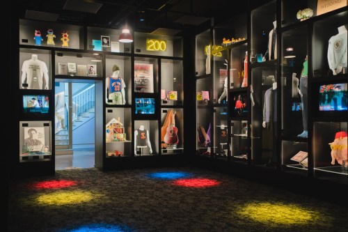 A dimly lit exhibit room showcasing various clothing items, memorabilia, and displays on black walls with colorful lights.