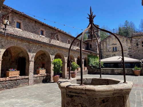 A stone courtyard with an ornate well, surrounded by rustic buildings and greenery under a clear blue sky.
