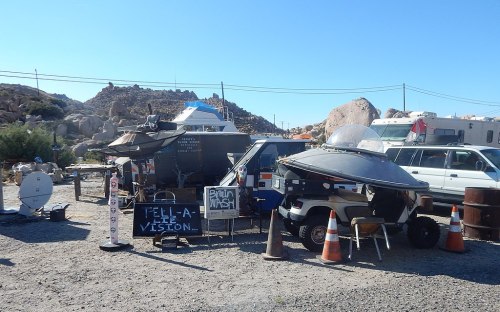 A quirky roadside setup with a sign for "TELL-A-LIE VISION," vehicles, and a car wash in a rocky desert landscape.