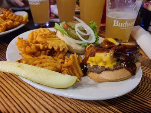 A plate with a cheeseburger topped with barbecue sauce, lettuce, pickles, and onion, served with waffle fries and a pickle slice.