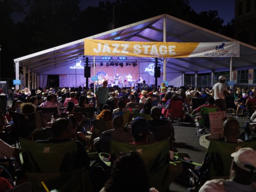 A large crowd enjoys a jazz performance under a tent at night, with a banner reading "Jazz Stage" overhead.