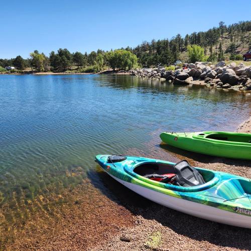 Two kayaks, one blue and one green, are parked on a sandy shore by a calm lake surrounded by trees and rocky terrain.