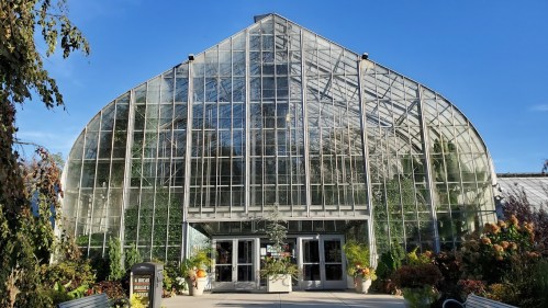 A large glass greenhouse with a curved roof, surrounded by greenery and colorful plants under a clear blue sky.