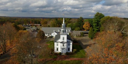 Aerial view of a white church with a steeple, surrounded by trees and autumn foliage under a cloudy sky.
