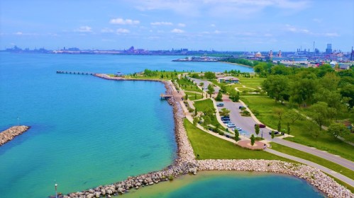 Aerial view of a serene lakeside park with green spaces, pathways, and industrial skyline in the background.