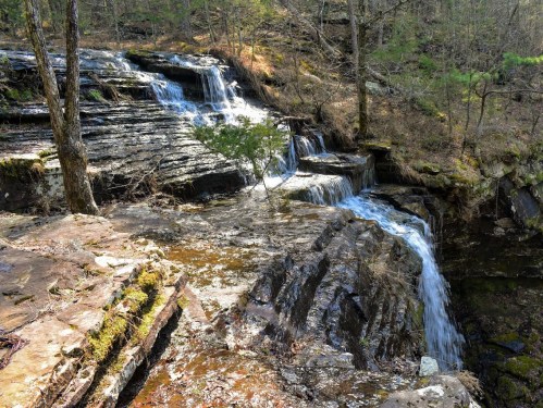 A serene waterfall cascades over rocky terrain, surrounded by trees and greenery in a peaceful forest setting.