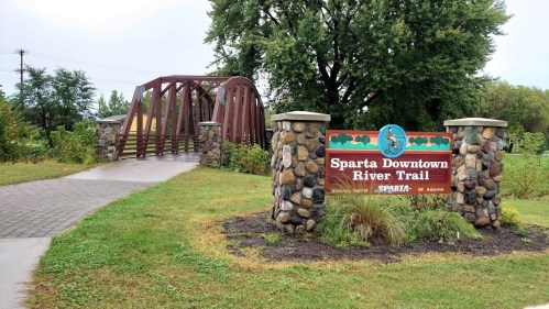 Sign for Sparta Downtown River Trail with a bridge in the background, surrounded by greenery and a paved pathway.