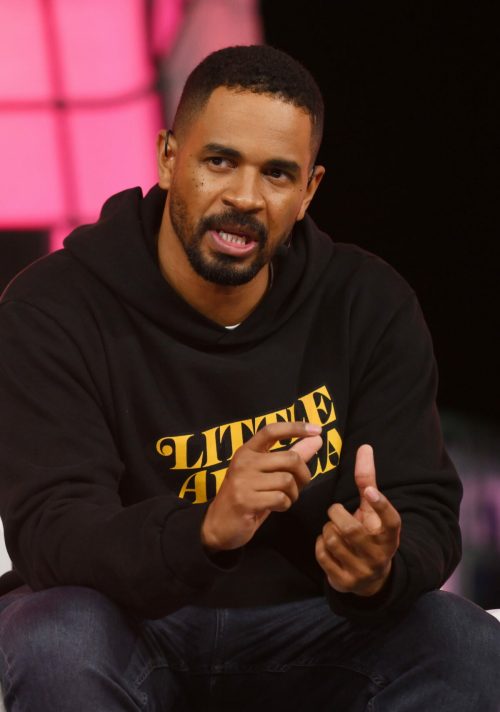 A man in a black hoodie speaks passionately at an event, gesturing with his hands.