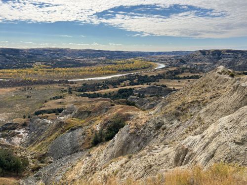 A scenic view of rolling hills and a winding river under a partly cloudy sky, with autumn foliage in the distance.