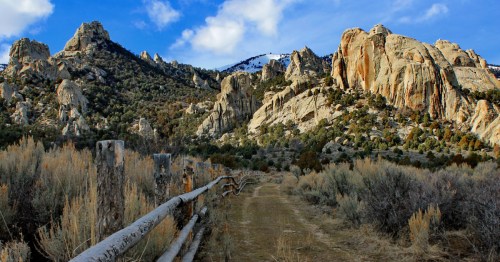 A dirt path leads through sagebrush, flanked by towering rocky cliffs under a blue sky with scattered clouds.