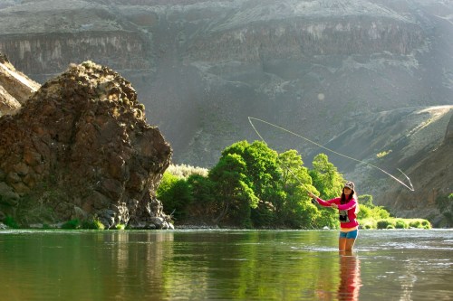 A person stands in a river, casting a fishing line with mountains and greenery in the background.