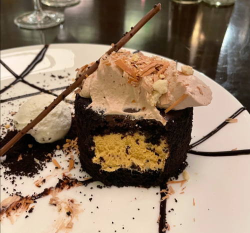 A slice of chocolate cake with creamy frosting, topped with chocolate shavings and a decorative stick, served with ice cream.