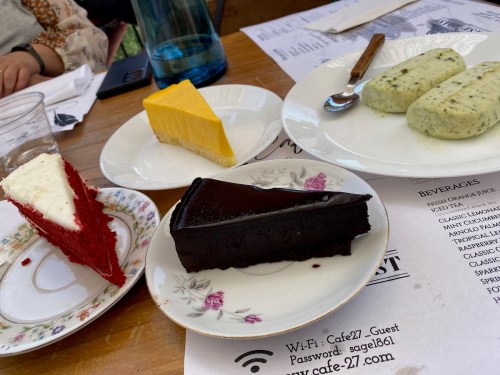 A table with four dessert slices: red velvet, lemon cheesecake, chocolate cake, and green tea mousse, alongside a menu.