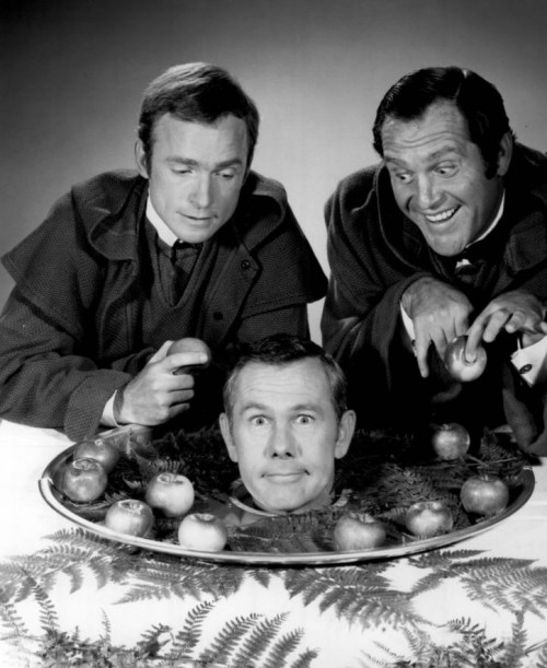 Two men in vintage attire humorously examine a platter with a man's head surrounded by apples. Black and white photo.
