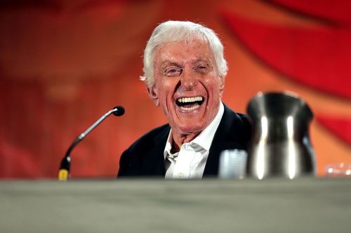 A smiling elderly man in a suit sits at a table, speaking into a microphone at an event.