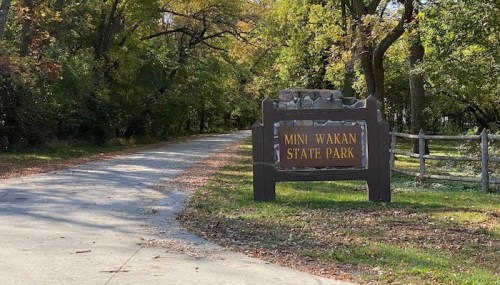Sign for Mini Wakan State Park at the entrance of a tree-lined road with autumn foliage.