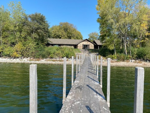 A wooden dock extends over calm water towards a stone house surrounded by trees under a clear blue sky.
