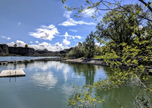 A serene lake surrounded by trees and a blue sky with fluffy clouds, reflecting the landscape on the water's surface.