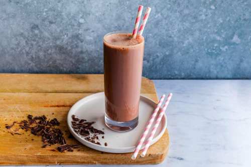 A tall glass of chocolate milkshake with two striped straws, served on a plate with chocolate shavings nearby.