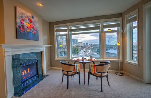 Cozy dining area with a fireplace and large windows overlooking a cityscape and waterfront.